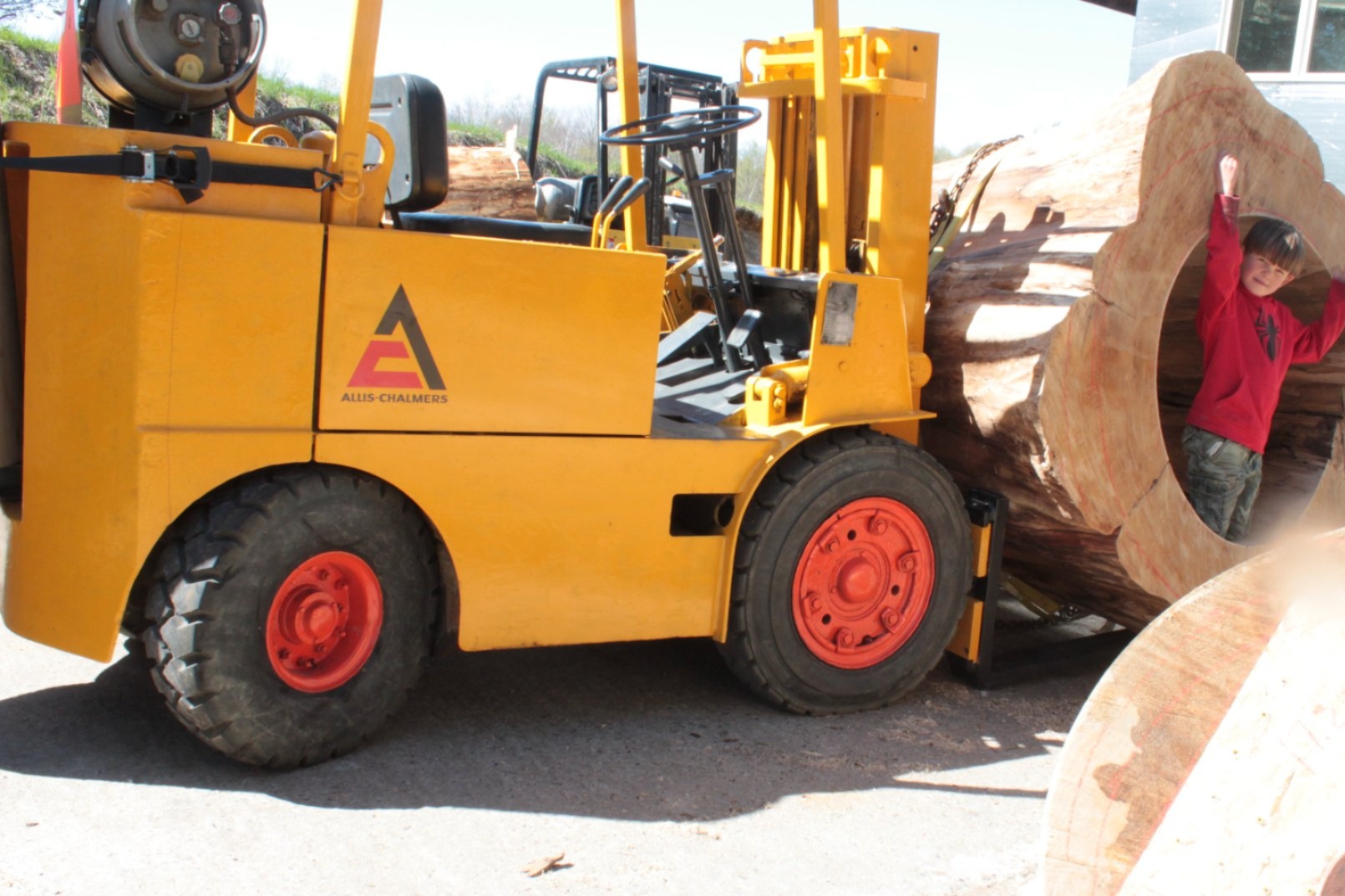 Boy waving from inside a massive IC WOOD hollow log cross-section leaning against the yellow Allis-Chalmers forklift — second large log cross-section in foreground, full production yard visible